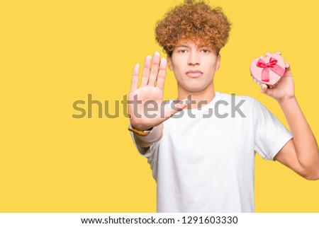 Young handsome man holding heart box as gift of valentine's day with open hand doing stop sign with serious and confident expression, defense gesture