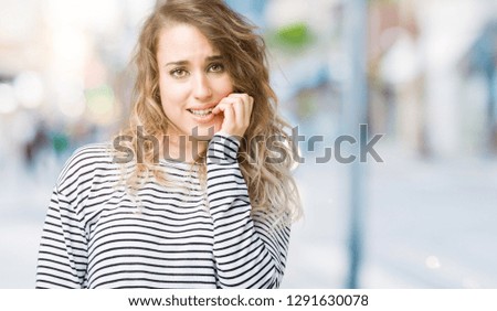 Beautiful young blonde woman wearing stripes sweater over isolated background looking stressed and nervous with hands on mouth biting nails. Anxiety problem.