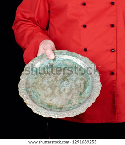 chef in red uniform holds in his hand an empty iron round dish, black background