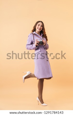 Full length photo of elegant woman 20s with long hair smiling and holding retro camera standing isolated over beige background