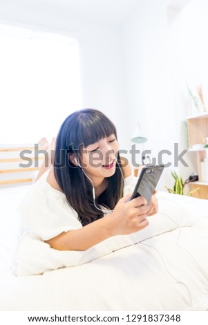 An Asian woman lies prone on her bed using her mobile phone and listening to music. This is the Internet of Things. This is lifestyle.

