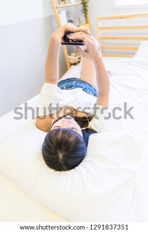 An Asian woman lies prone on her bed using her mobile phone and listening to music. This is the Internet of Things. This is lifestyle.

