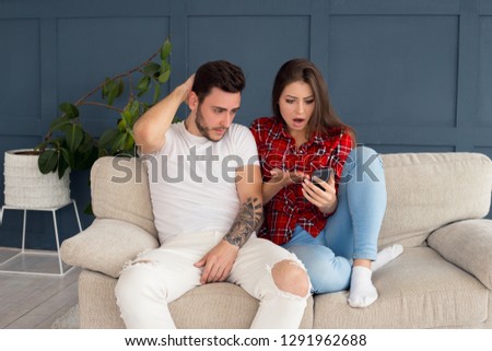 Bad news! Shocked frustrated young couple sitting on the sofa and disappointing the news, they having seen on the screen of the smartphone.