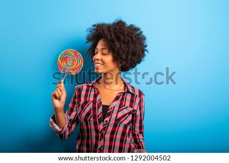 Smiling curly african girl in bright yellow dress holding candy lollipop. Blue studio background, copy space. Sweet life and confectionary - Image