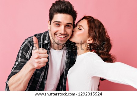 Charming ginger girl kissing husband on pink background. Happy couple making selfie together.