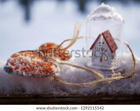 A small white toy house with a red roof and a lighted illumination and a pair of orange felt hearts on the snow covered with snow, the concept of winter seasonal holidays, Valentine's Day greetings, h
