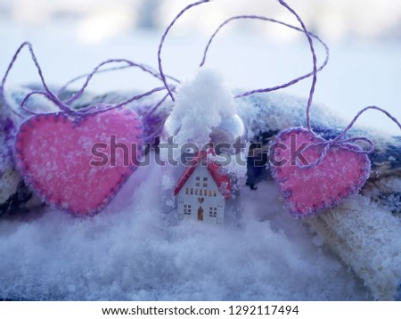 A small white toy house with a red roof and a lighted illumination and a pair of pink felt hearts on the snow covered with snow, the concept of winter seasonal holidays, Valentine's Day greetings, hom