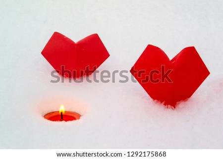 Two paper hearts (origami) and a burning tea candle are set in the snow. Picture for Valentine's Day.