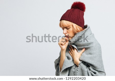 Cute sick young teen woman in red hat, wrapped in warm scarf coughing, closed eyes.Female feeling the first symptoms of illness.Bronchitis, upper respiratory infection concept.Moist cough, laryngitis Royalty-Free Stock Photo #1292203420