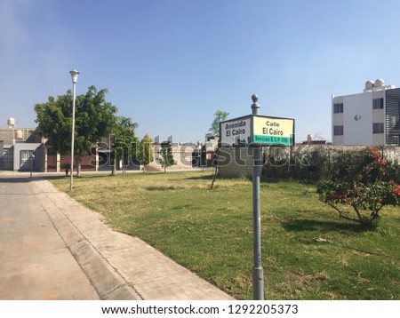 Mexican neighborhood street sign to the right with a grassy garden and bush in the middle and buildings in the background and rubbish at the bottom.