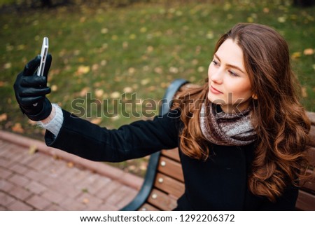 Young cute brunette with short hair is sitting on wooden bench, making selfie with kissing lips on her smartphone in a black coat,girl in the park with a phone,woman is relaxing outdoors