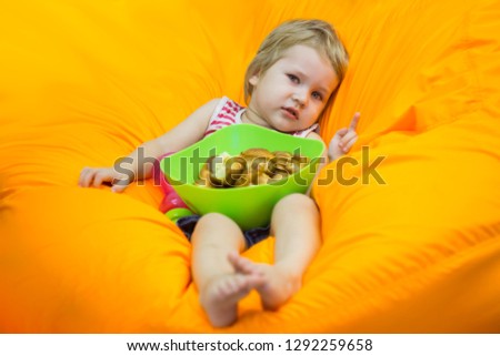 A little boy wallows in an orange bean bag chair with a green basin of simple cupcakes and watches cartoons.