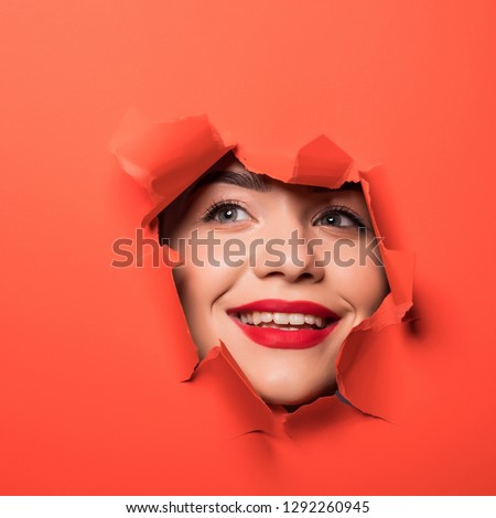 The face of young beautiful girl with a bright make-up and with plump red lips peeks into a hole in orange paper