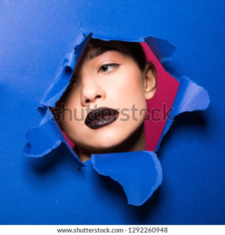 The face of a young beautiful girl with a bright make-up and puffy blue lips peers into a hole in blue paper.