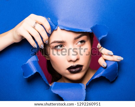 The face of a young beautiful girl with bright make-up and puffy dark lips peers into a hole in blue paper.