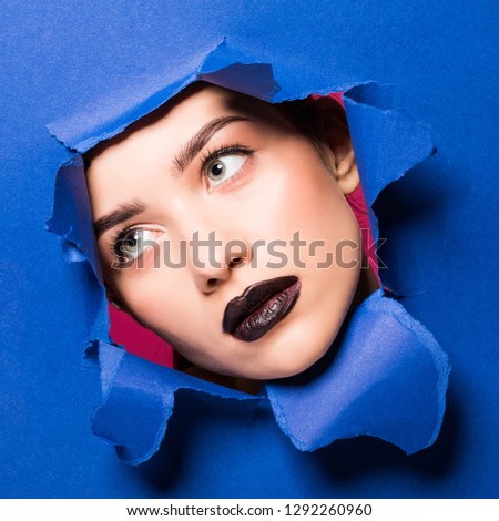The face of a young beautiful girl with a bright make-up and puffy blue lips peers into a hole in blue paper.