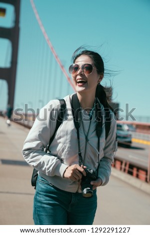 cheerful tourist photographer laughing holding professional camera standing under sunshine on red golden gate bridge in san francisco on pedestrian with cars dirving pass on road in background.