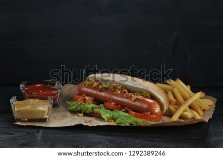 Hotdog and french fries on crayfish paper. Hotdog stuffing consists of sausage, lettuce, tomato slices, cucumber salad, dried onion, mustard and ketchup. Close-up. Dark background.