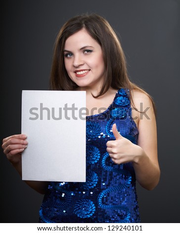 Happy young woman in a blue dress. Woman holds a poster in her right hand. Left hand showing thumbs up. On a gray background