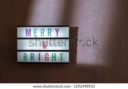 Message Merry & Bright on illuminated board. Holidays concept with text. Daylight from window. Room interior. Coloured letters Merry and Bright on purple wallpaper wall.