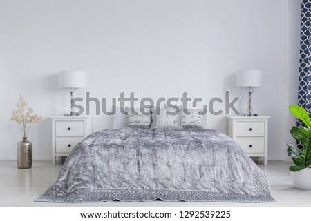 Grey bedding on king size bed in stylish bright bedroom interior with white bedside tables and lamps, real photo with copy space on empty white wall