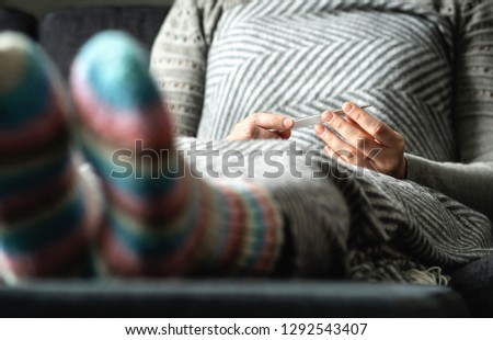 Sick woman with flu and cold checking fever and body temperature with thermometer. Ill person sitting on couch at home under blanket with warm woolen socks. Virus, infection, norovirus or hypothermia. Royalty-Free Stock Photo #1292543407