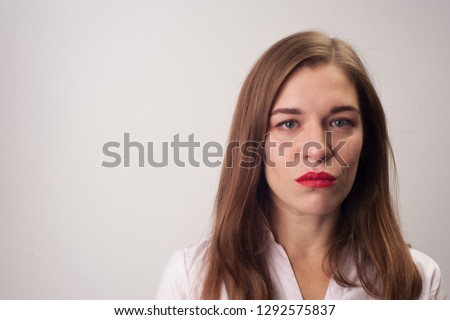 Portrait close-up of sad upset woman blue eyes with light-brown hair loosed and dressed in white blouse smiling sadly looking at the camera standing on white background.