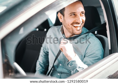 Portrait of smiling Caucasian man fastening seat belt and sitting in his car. Window opened, side view. Royalty-Free Stock Photo #1292580019