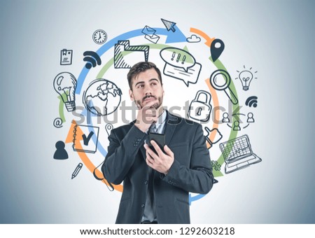 Portrait of thoughtful young businessman with beard wearing suit and holding smartphone standing near gray wall with internet icons drawn on it.