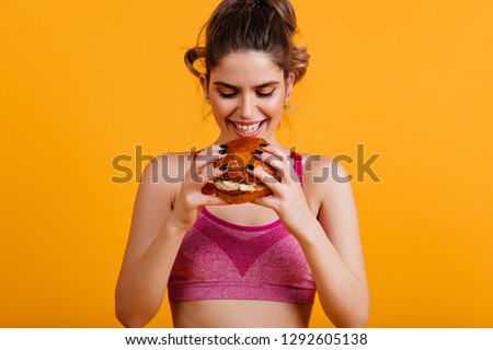 Smiling caucasian girl eats burger. Indoor portrait of amazing woman with sandwich.