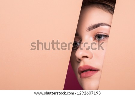 The face of young beautiful girl with a bright make-up and puffy orange lips peers into a hole in peach paper.