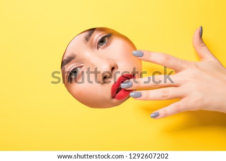 A girl with beautiful long fingers and red nails touches the edge of her eye and looks into the circular hole of the yellow paper.