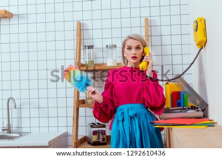 surprised housewife in colorful clothes holding dusting brush and talking on retro telephone in kitchen