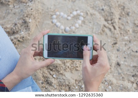 female hands with smartphones take pictures of shells on the sand in the form of a heart