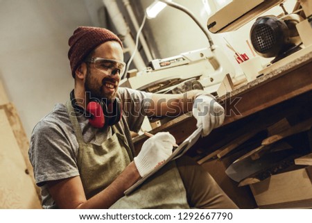 Attractive male in apron and safety goggles cheerfully smiling and drawing sketches in sketchbook while sitting near workbench in professional workshop