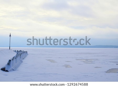 A view of a frozen lake covered in snow and ice and with some trees visible next to cloudy horizon with some concrete trapezoids and a lamp near the coast seen during a cold winter day in Poland