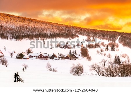 Wonderful view of the calm, snowy and landscape, hillside with beautiful mountain cottages, alleys, road and forest, silhouette of people with a stroller and golden cloudy sky in the background