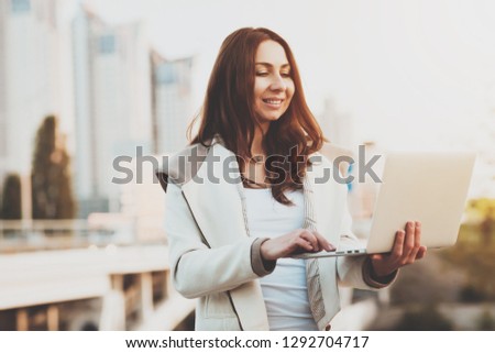 A young girl looks at information on a laptop. Young smiling beautiful girl looking information on a laptop while standing outdoors. Technologies helping in work. The concept business person.