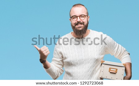Young hipster man wearing glasses, moving holding moving box over isolated background pointing and showing with thumb up to the side with happy face smiling