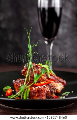 The concept of American cuisine. Roasted pork ribs, baked and glazed in barbecue sauce. Serving dishes in the restaurant on a black plate with sesame and micro green. Next to a glass of red wine.