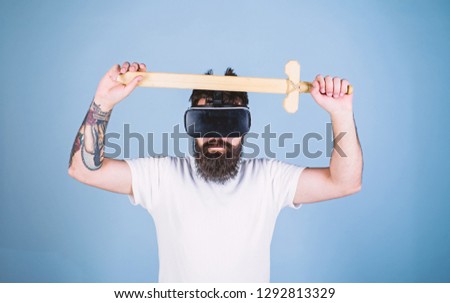 Gamer concept. Man with beard in VR glasses, light blue background. Guy with head mounted display holds sword, play fighting game in VR. Hipster on serious face enjoy play game in virtual reality.