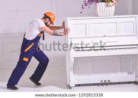 Heavy loads concept. Loader moves piano instrument. Courier delivers furniture, move out, relocation. Man with beard worker in helmet and overalls pushes, put efforts to move piano, white background.