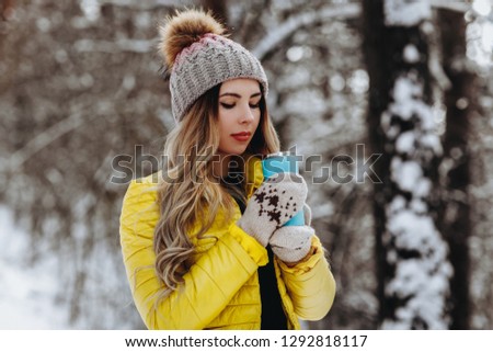 beautiful girl standing in winter forest and drink hot beverage. Wonam wear yellow coat, knitted gloves and hat with ball. Beauty long blond lush hair. winter holiday concept.