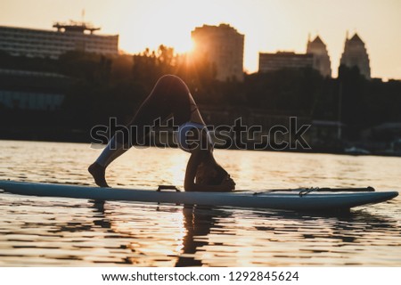 Flexible girl stretching muscles in downward facing dog asana. She standing on inflatable board on river with city silhouette on background