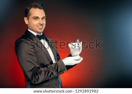 Professional magician wearing suit and gloves standing isolated on blue and red background showing tricks shuffling cards looking camera smiling playful