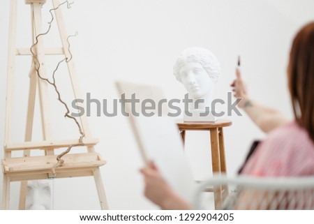 Young woman artist draws a pencil on canvas. White studio, pink shirt and apron. Drawing and painting lessons, professional artist.