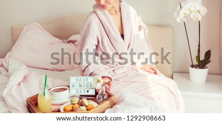 Good morning concept. Woman in bathrobe sitting on the bed and has breakfast tray with coffee, macaroons and Have a nice day text on lighted box. Hospitality, care, service. Wide banner. Copy space