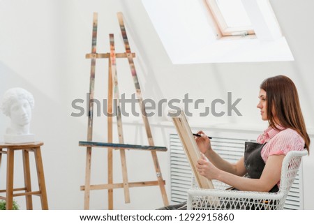Young woman artist draws a pencil on canvas. White studio, pink shirt and apron. Drawing and painting lessons, professional artist.
