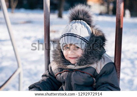 A cute boy in winter clothes and in warm gray hats with a hood on his head skates in the winter on a swing and smiles. Portrait of a beautiful boy with blue eyes in winter