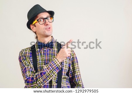Funny nerd pointing finger away at copy space. Smiling young nerdy in glasses showing your product. Nerd man in plaid shirt and suspender, isolated on white background. Excited young guy presentation